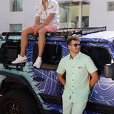 Man leaning against a vehicle wearing the Baja Llama Go Fast Green Vagabond Button Up shirt and matching shorts