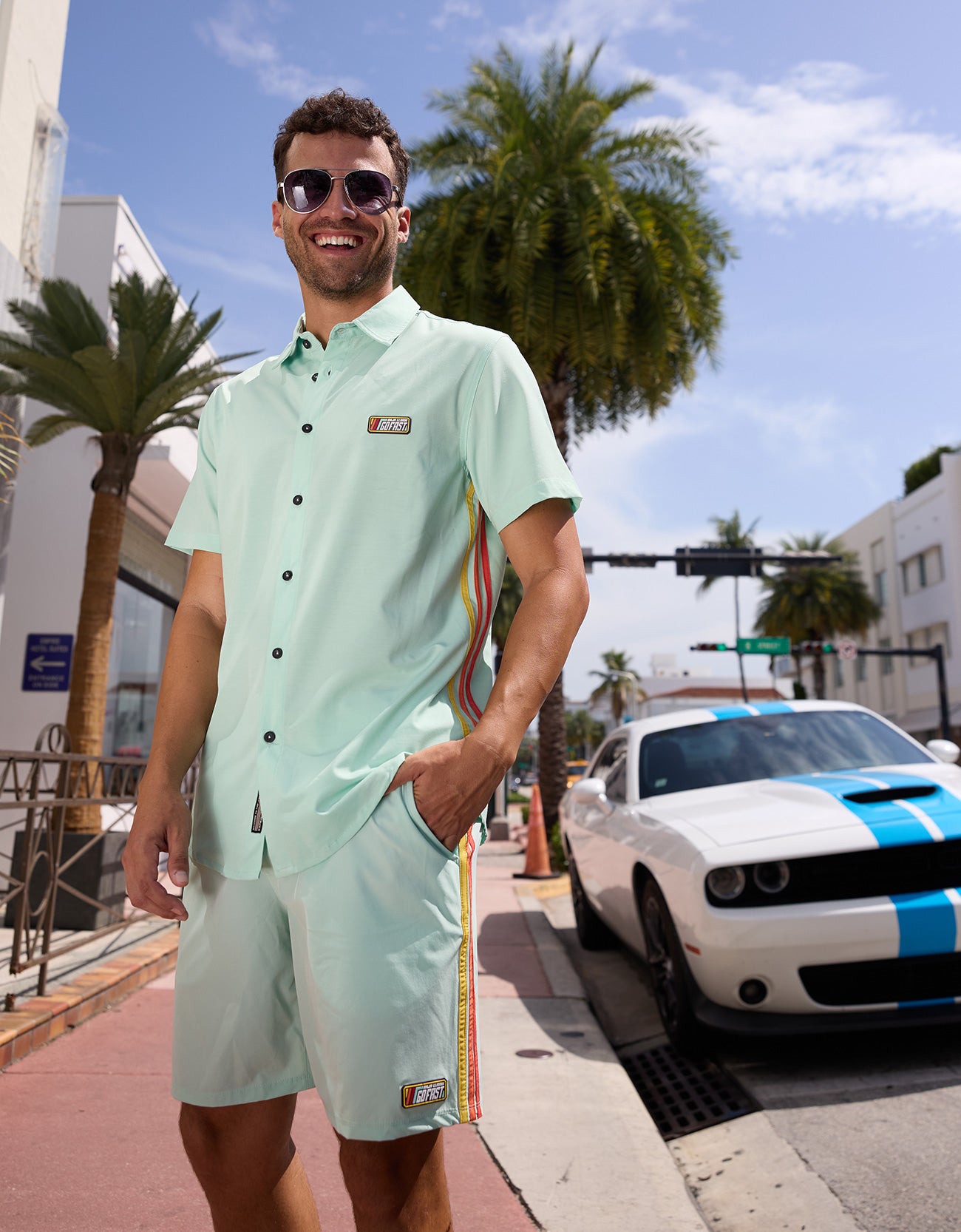 Man wearing Baja Llama Go Fast Green Vagabond Button Up shirt and matching shorts outdoors with a sports car