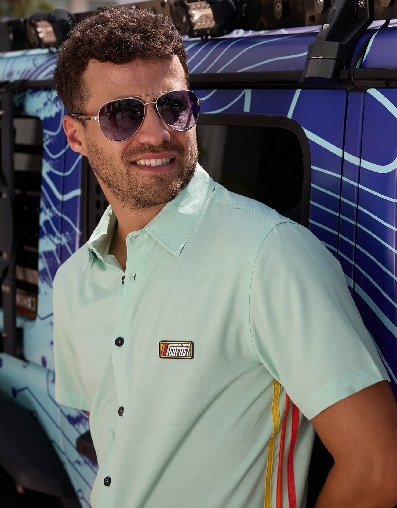Baja Llama Go Fast Green Vagabond Button Up shirt worn by a man wearing sunglasses next to a vehicle