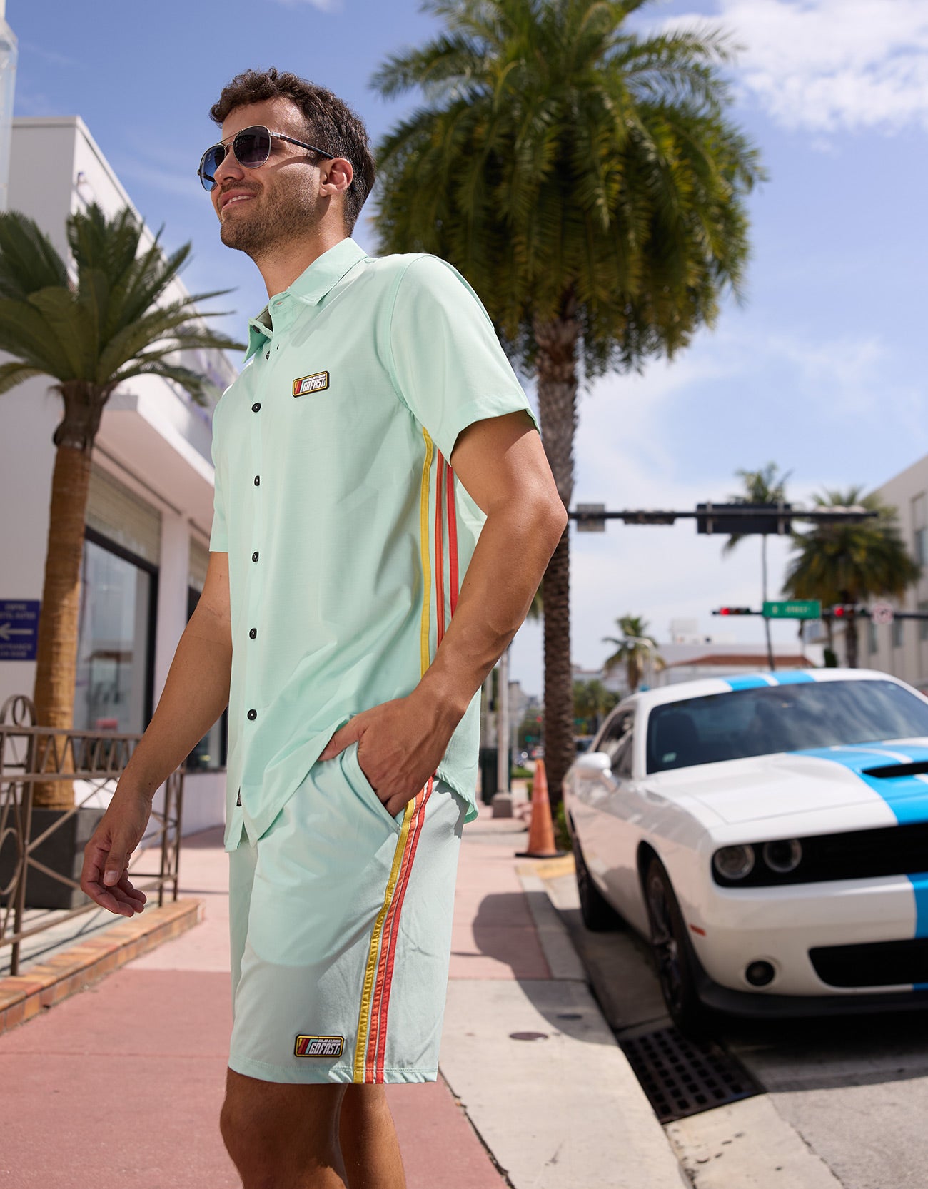 Man wearing the Baja Llama Go Fast Green Vagabond Button Up shirt and matching shorts outdoors by a sports car