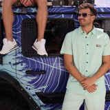Man wearing the Baja Llama Go Fast Green Vagabond Button Up shirt and matching shorts standing beside a patterned vehicle