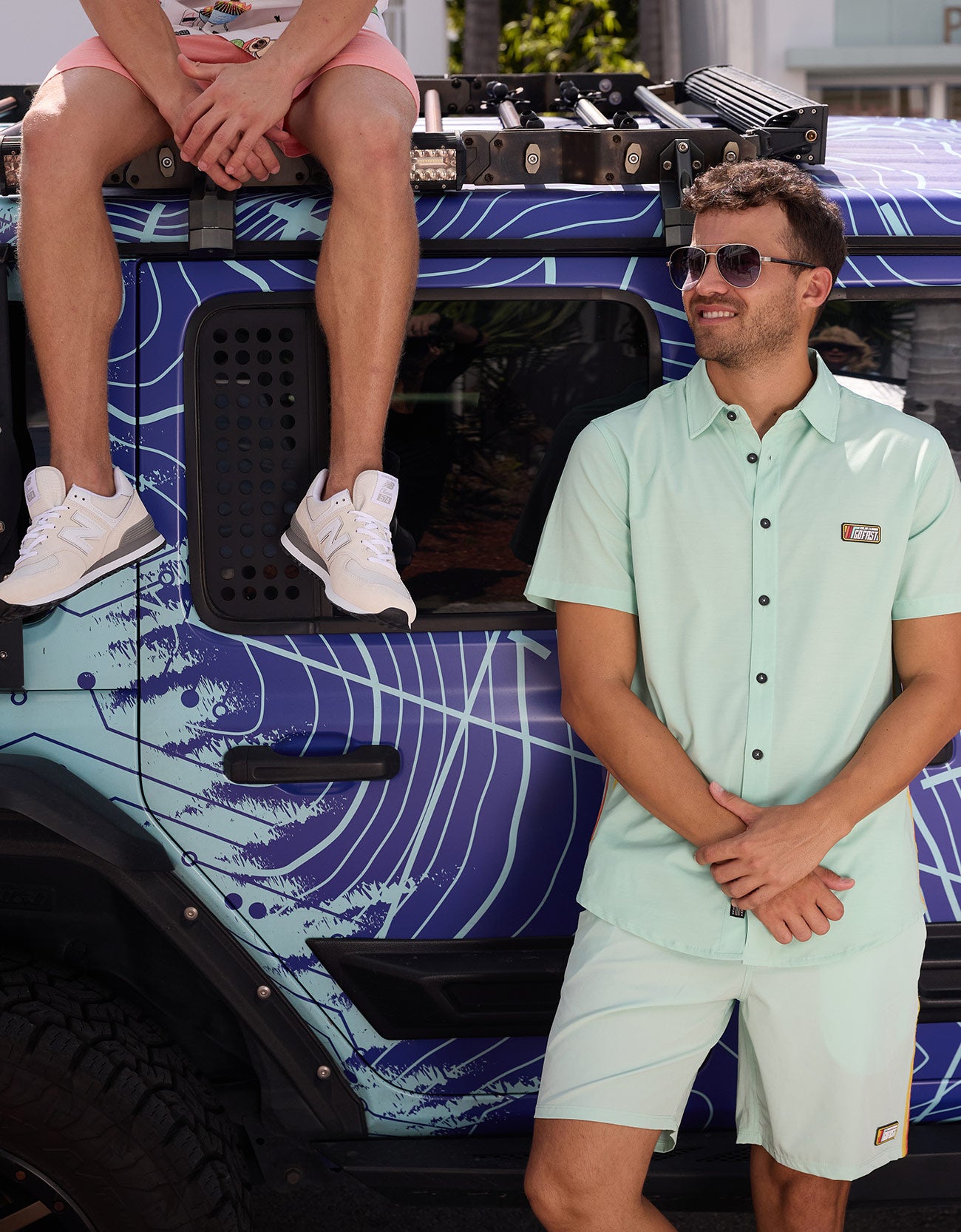 Man wearing the Baja Llama Go Fast Green Vagabond Button Up shirt and matching shorts standing beside a patterned vehicle