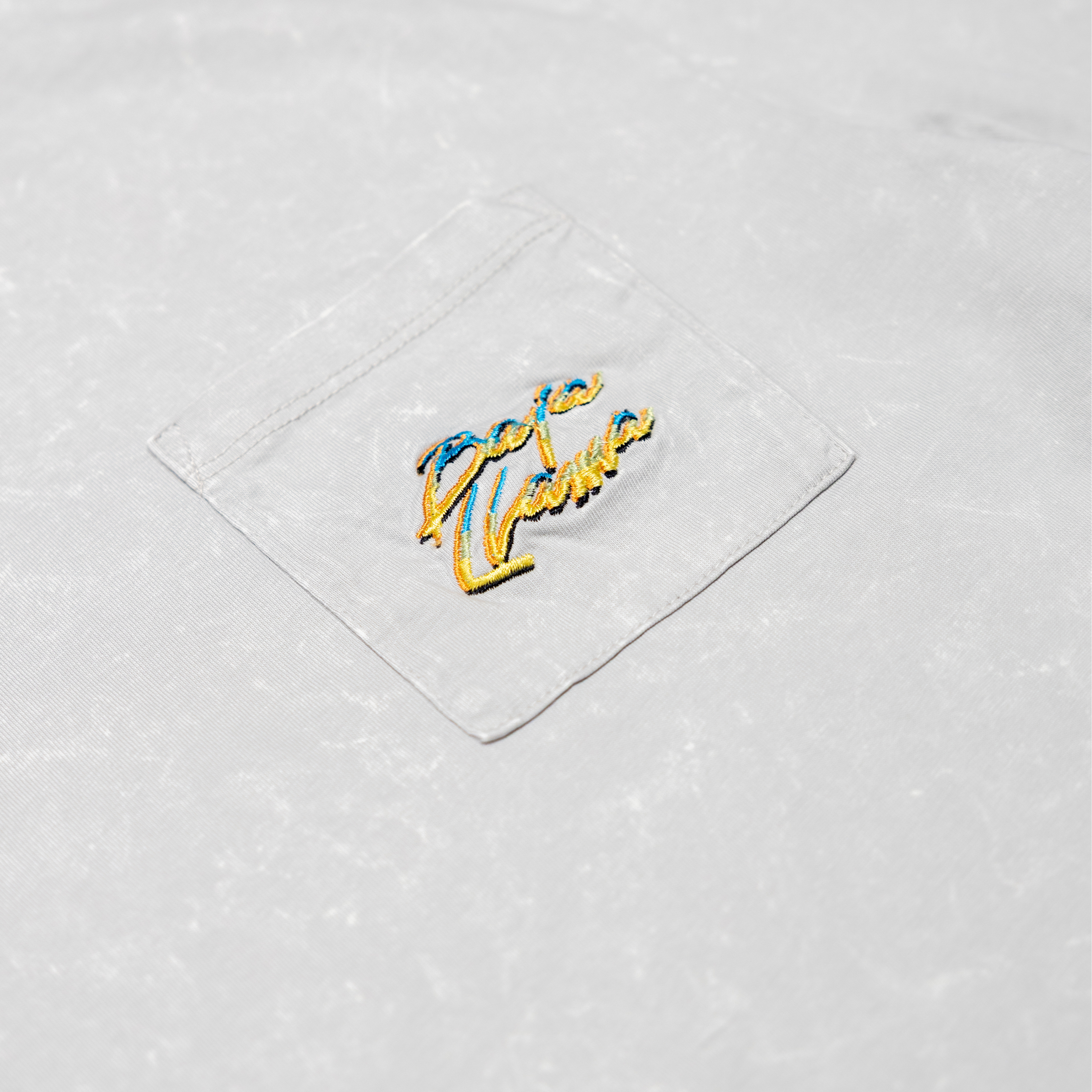 Close-up of the front chest pocket on the Baja Llama Greatful Dyed Shoreline Smoke Heavy Tee, showing the yellow and blue embroidered cursive brand logo.