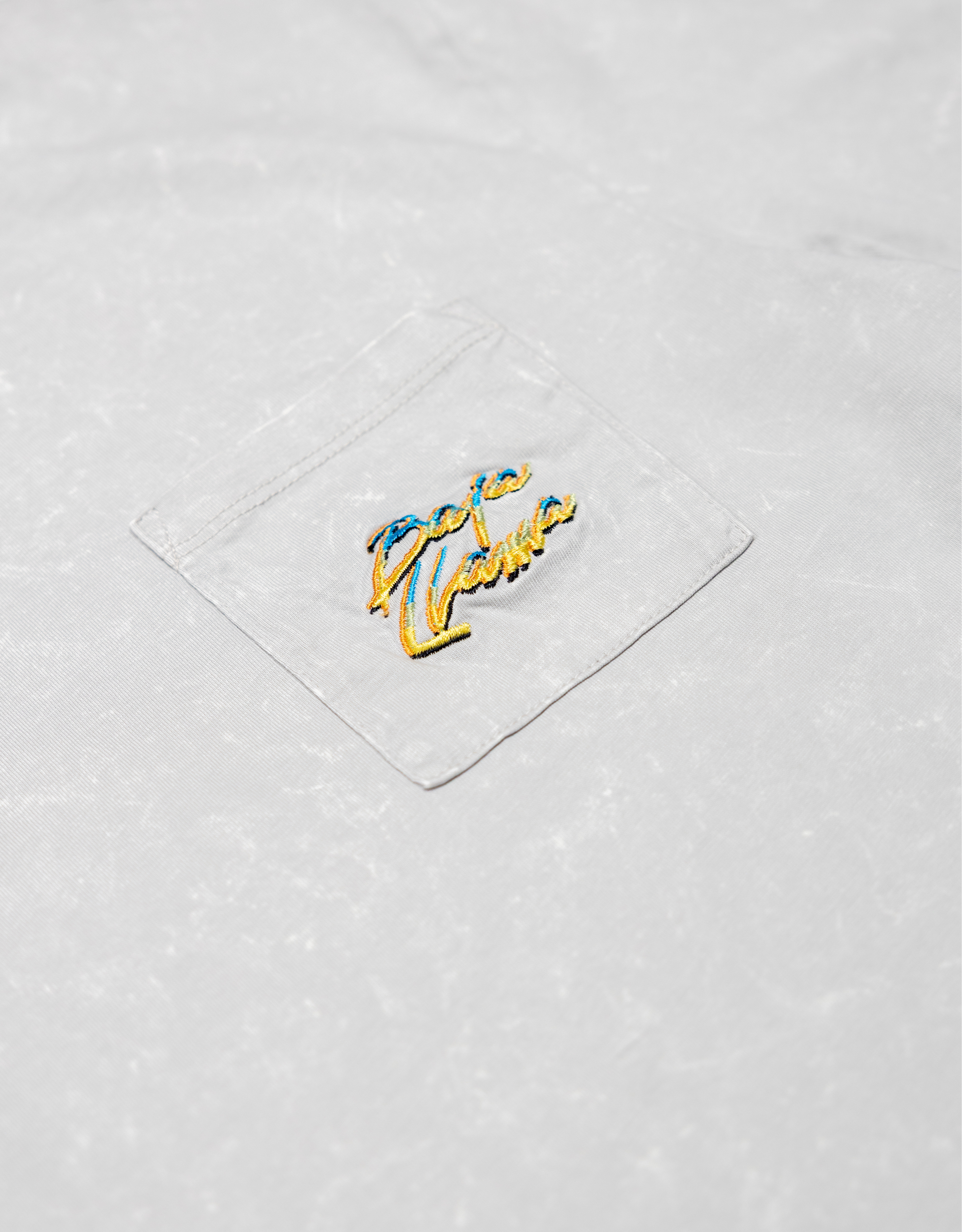 Close-up of the front chest pocket on the Baja Llama Greatful Dyed Shoreline Smoke Heavy Tee, showing the yellow and blue embroidered cursive brand logo.