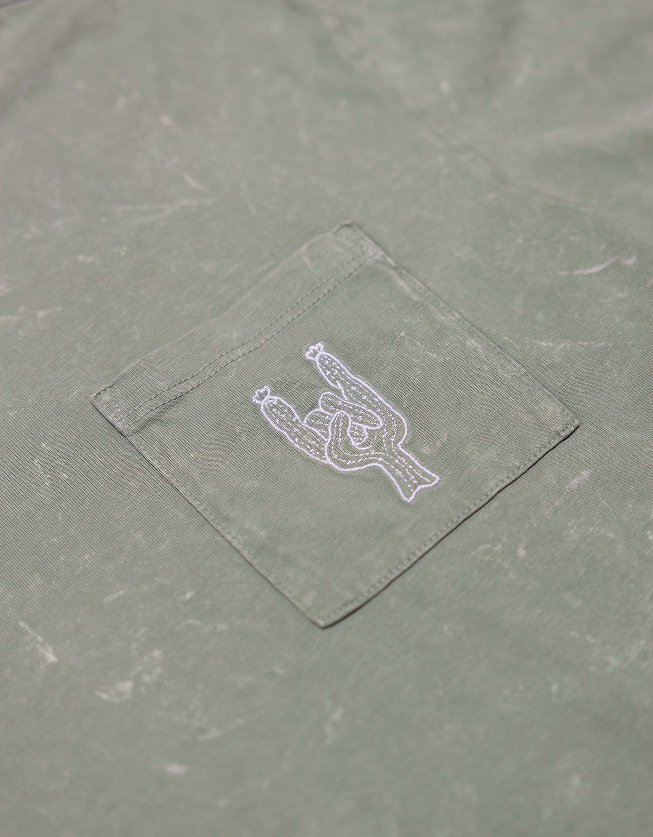 Close-up of the front chest pocket on the Baja Llama Greatful Dyed Rock On Cypress Green Heavy Tee, showing a white embroidered cactus in the shape of a rock-on sign