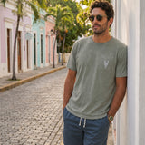 Man leaning against a white wall wearing the Baja Llama Greatful Dyed - Rock On Cypress Green Heavy Tee on a cobblestone street