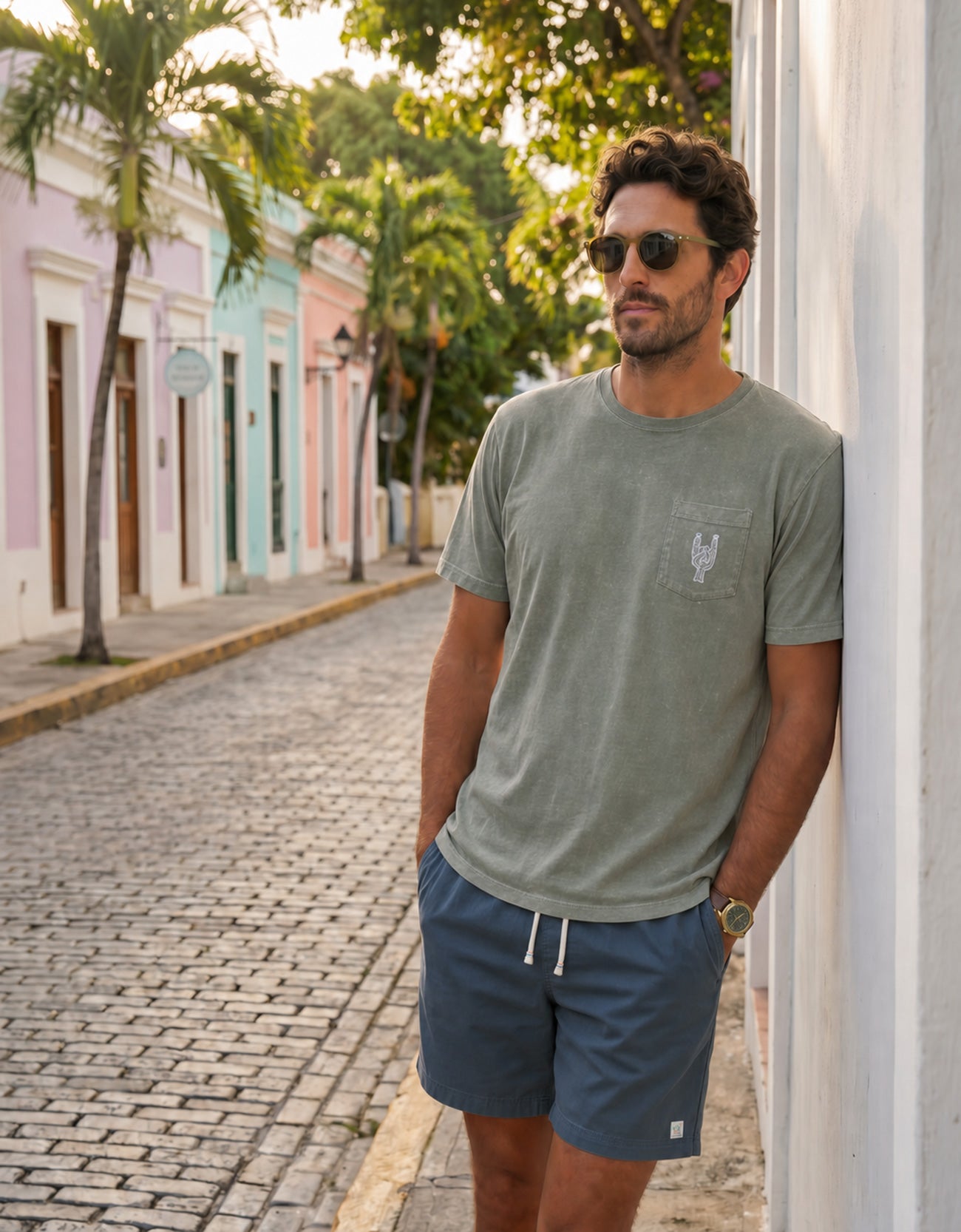 Man leaning against a white wall wearing the Baja Llama Greatful Dyed - Rock On Cypress Green Heavy Tee on a cobblestone street