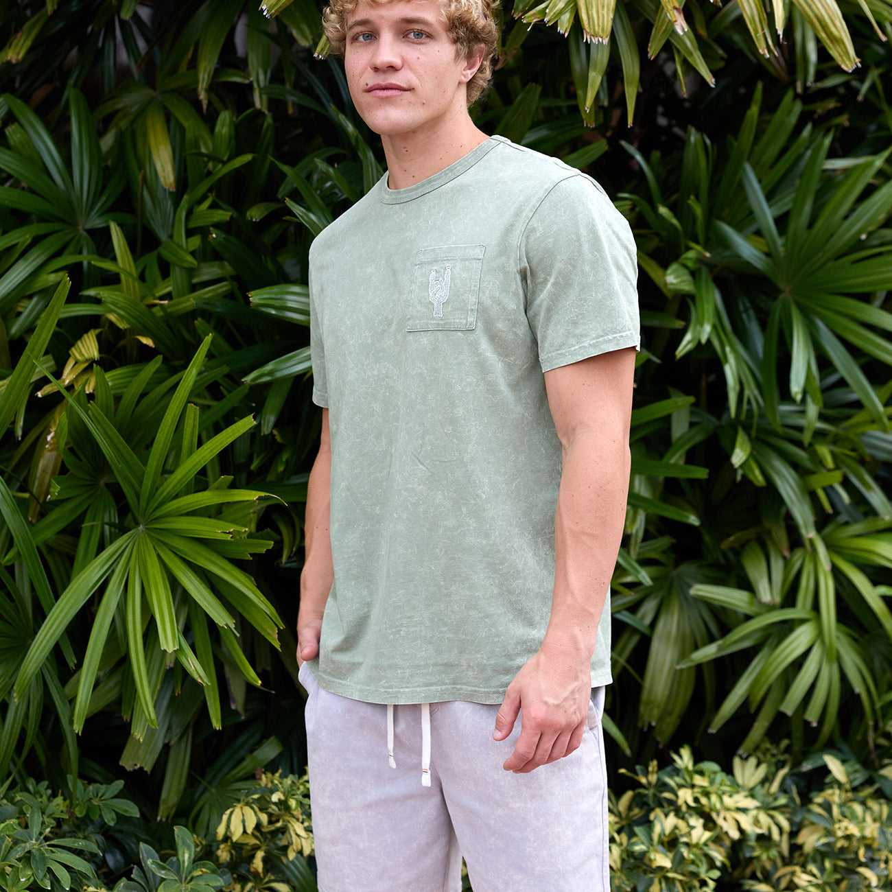 Man wearing the Baja Llama Greatful Dyed - Rock On Cypress Green Heavy Tee outdoors among tropical foliage