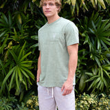 Man wearing the Baja Llama Greatful Dyed - Rock On Cypress Green Heavy Tee outdoors among tropical foliage