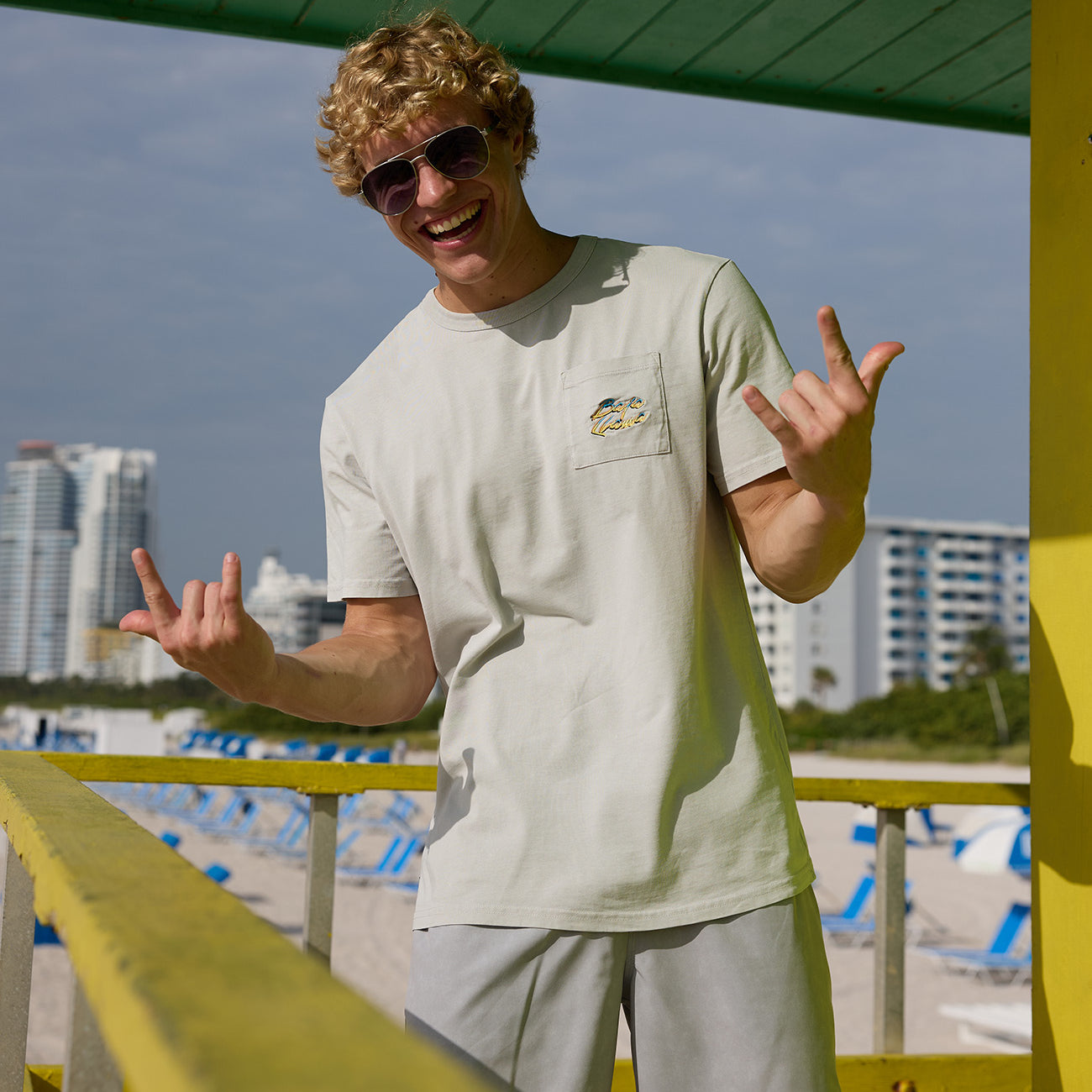 Man flashing hand signs on a lifeguard tower wearing the Baja Llama Greatful Dyed - Shoreline Smoke Heavy Tee