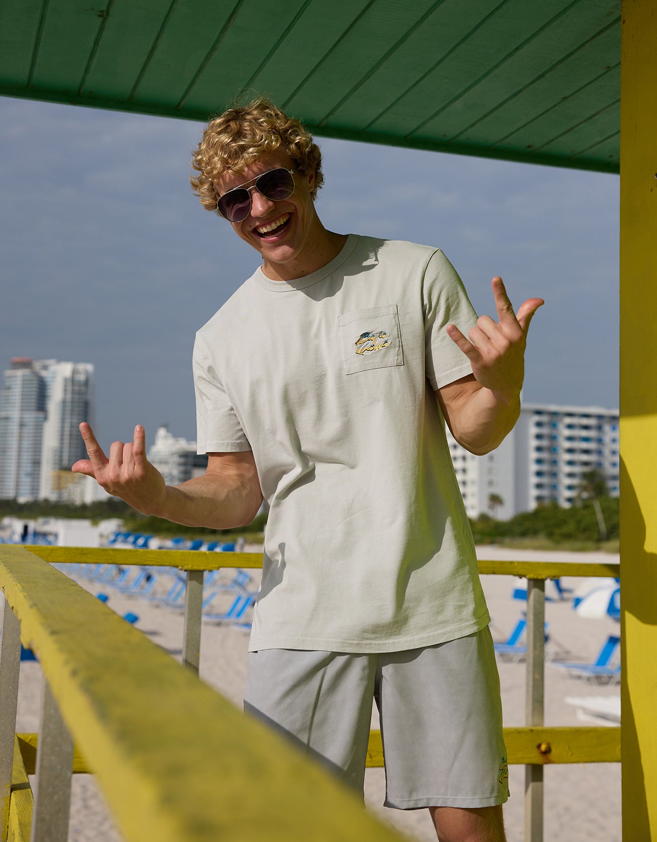 Man flashing hand signs on a lifeguard tower wearing the Baja Llama Greatful Dyed - Shoreline Smoke Heavy Tee