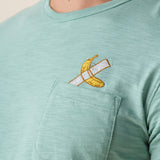 A close-up shot of the left chest on the Baja Llama La Banana Embroidered Pima Slub Pocket Tee as worn by a model, showing the textured sage green fabric  topped with a playful embroidered graphic of a banana fixed with white duct tape topped with a chest pocket