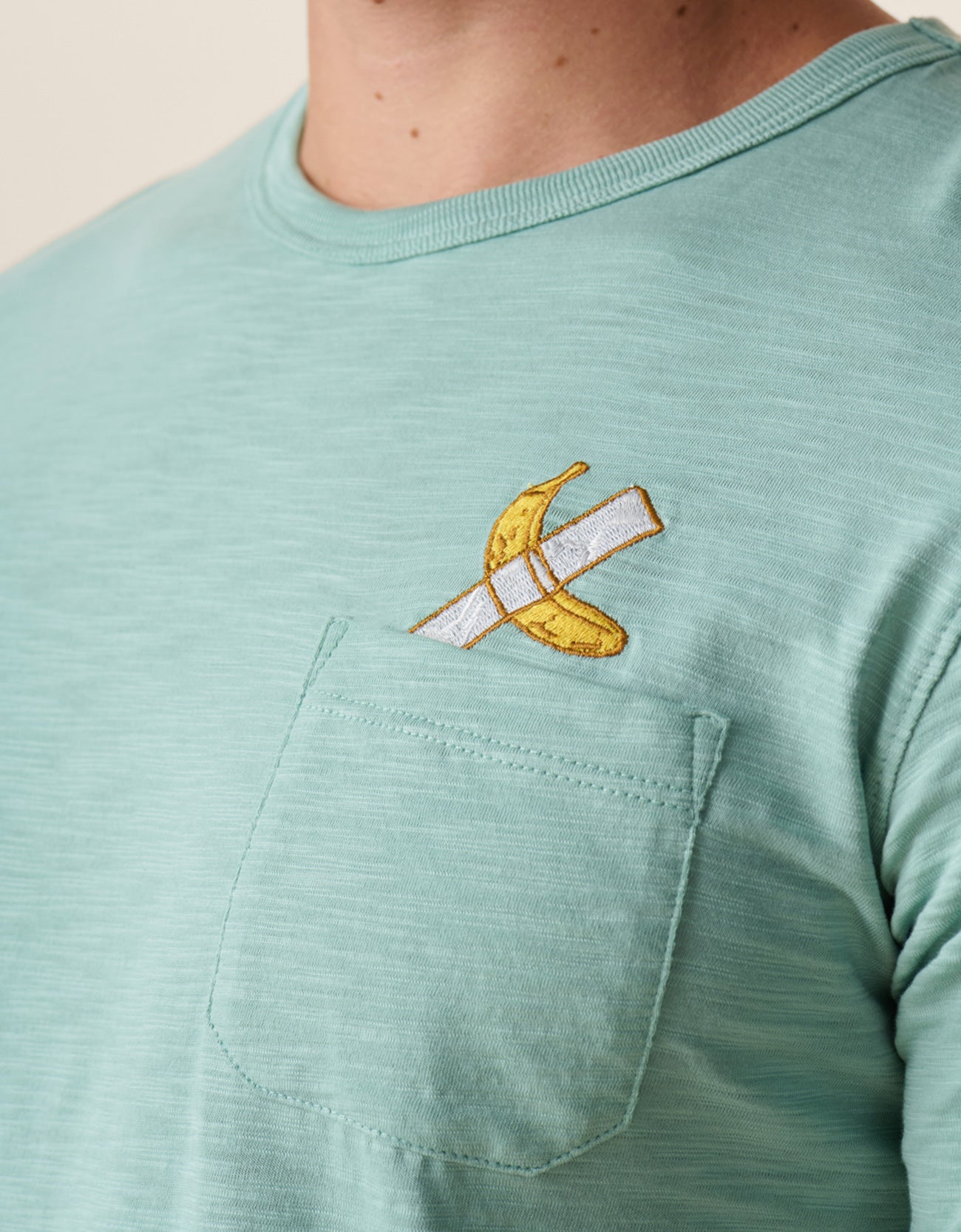 A close-up shot of the left chest on the Baja Llama La Banana Embroidered Pima Slub Pocket Tee as worn by a model, showing the textured sage green fabric  topped with a playful embroidered graphic of a banana fixed with white duct tape topped with a chest pocket
