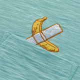 An extreme close-up shot of the left chest pocket area on the Baja Llama La Banana Embroidered Pima Slub Pocket Tee, showcasing the textured sage green fabric and a detailed embroidery of a yellow banana fixed with white duct tape.