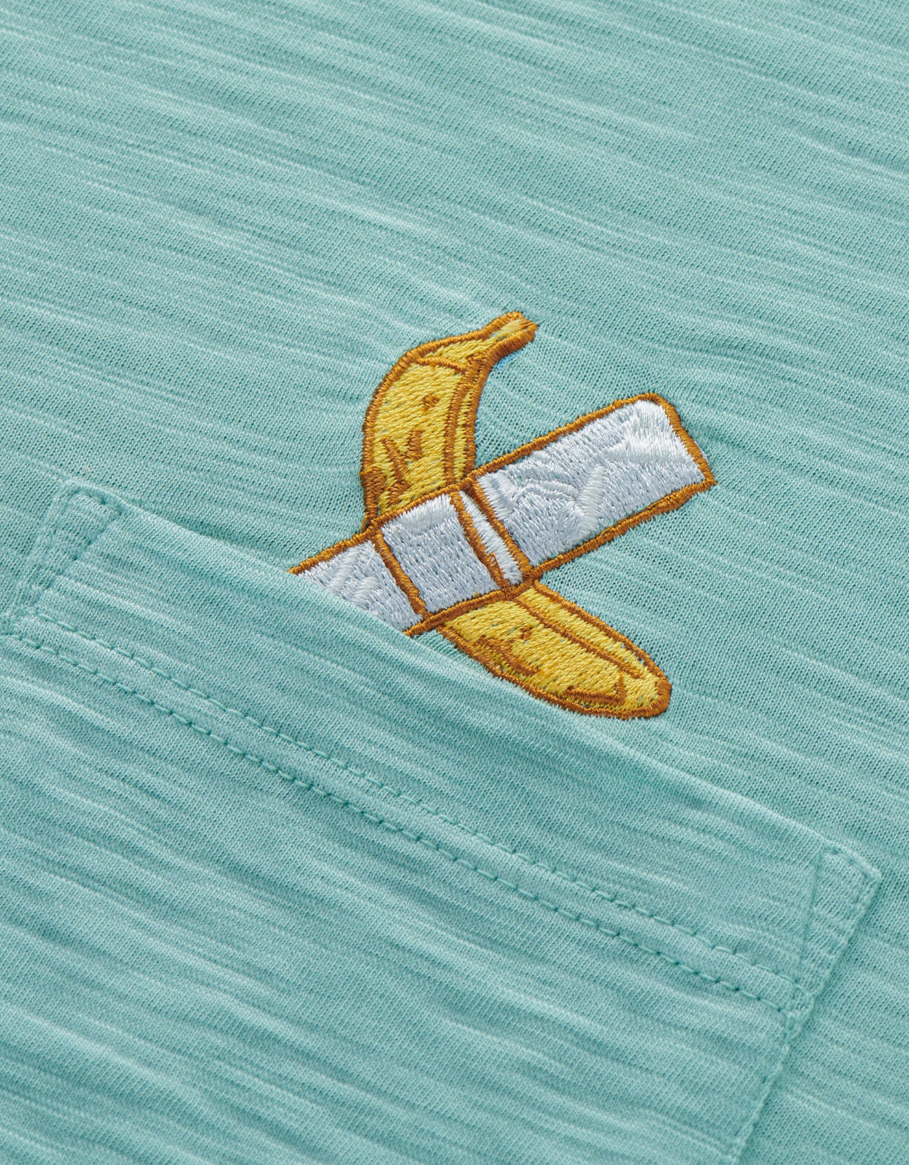 An extreme close-up shot of the left chest pocket area on the Baja Llama La Banana Embroidered Pima Slub Pocket Tee, showcasing the textured sage green fabric and a detailed embroidery of a yellow banana fixed with white duct tape.