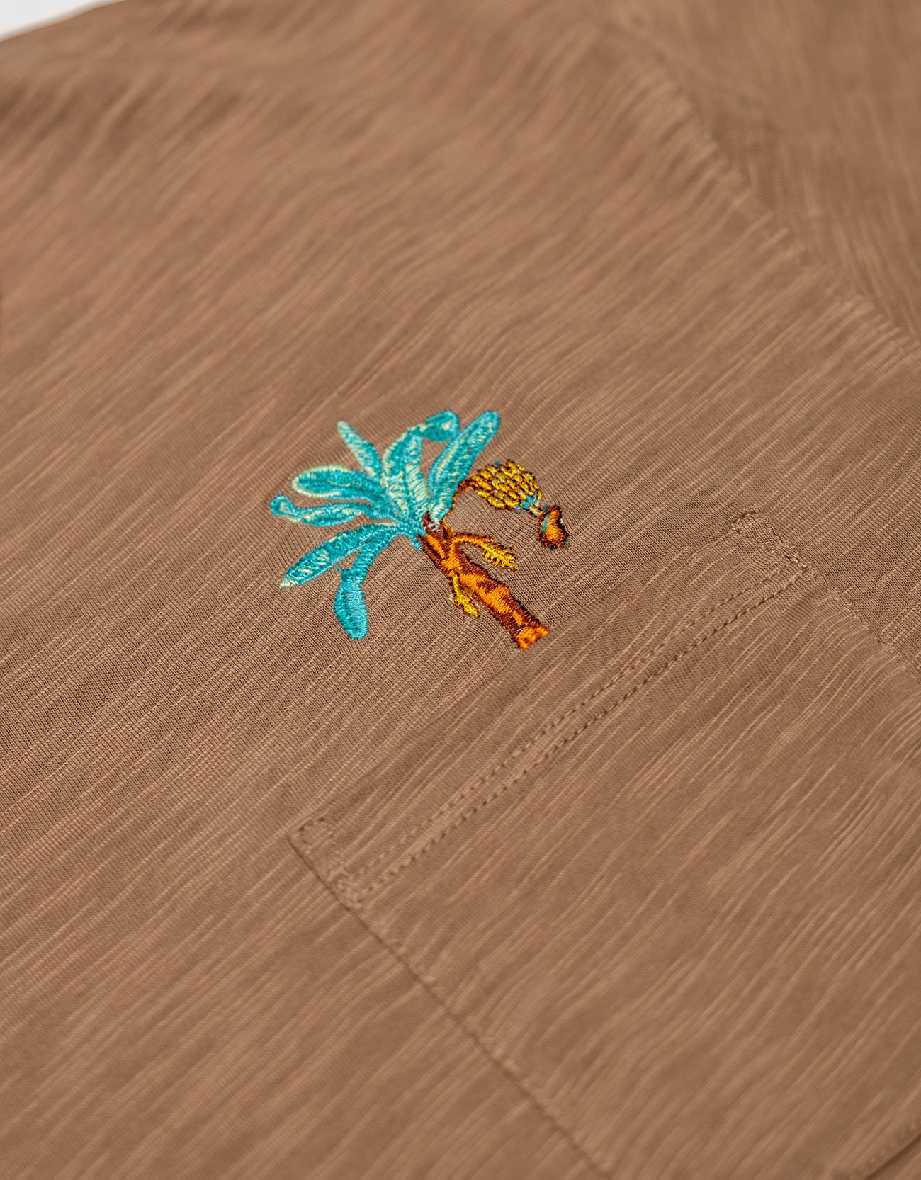 An extreme close-up shot of the left chest pocket area on the Baja Llama La Republica Embroidered Pima Slub Pocket Tee, highlighting the detailed brown slub fabric texture and an embroidered turquoise and orange banana tree with a hanging bunch of bananas.