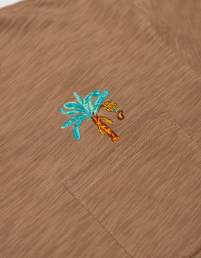 An extreme close-up shot of the left chest pocket area on the Baja Llama La Republica Embroidered Pima Slub Pocket Tee, highlighting the detailed brown slub fabric texture and an embroidered turquoise and orange banana tree with a hanging bunch of bananas.