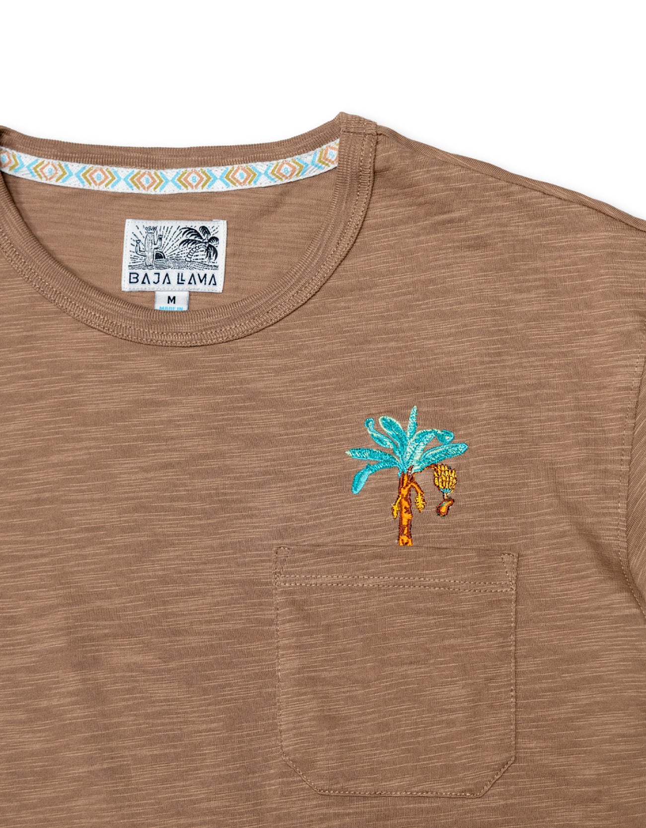 A close-up shot of the neckline and chest area on the Baja Llama La Republica Embroidered Pima Slub Pocket Tee, showing the brown textured fabric, a stitched pocket with an embroidered teal banana tree, and a geometric pattern on the inner collar.