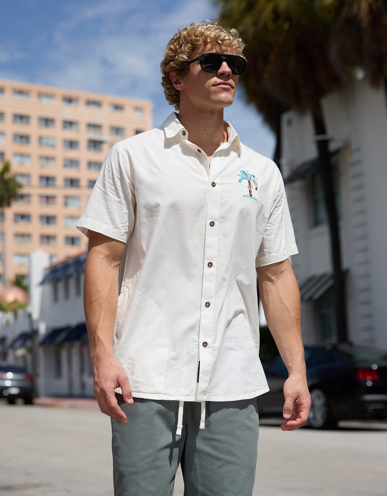 Model walking outdoors wearing Baja Llama La Republica Embroidery 7-Seas Relaxed Fit Button Up with palm tree embroidery and sunglasses