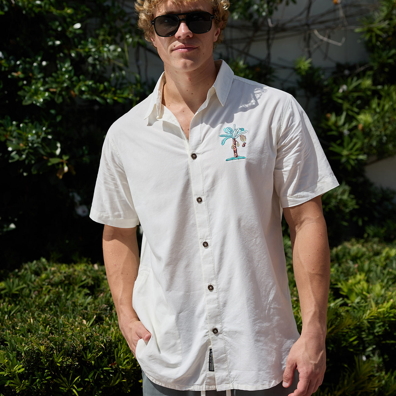 Man wearing white short-sleeve button-up shirt with embroidered palm tree pocket design outdoors