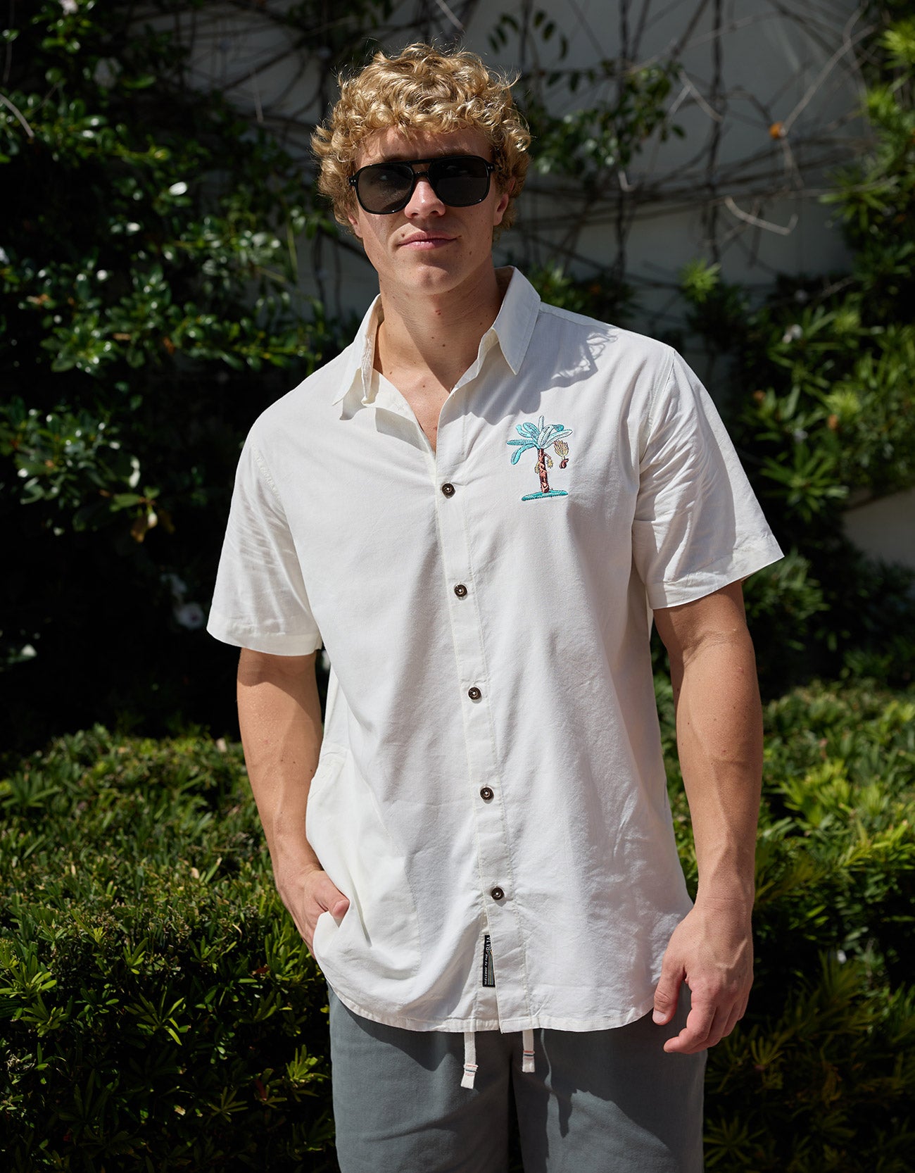 Man wearing white short-sleeve button-up shirt with embroidered palm tree pocket design outdoors