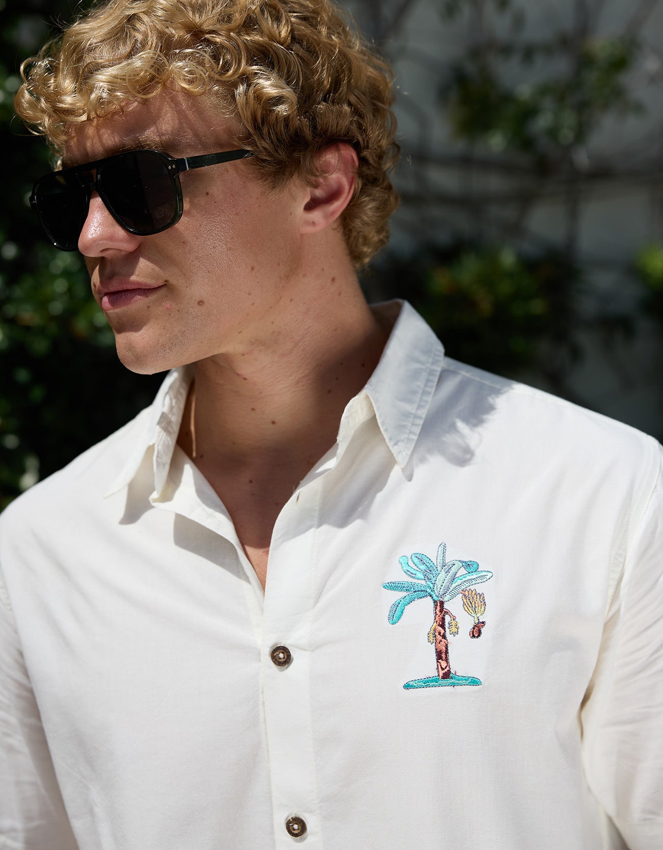 Close-up of model wearing white 7-Seas button-up shirt featuring embroidered chest palm tree design and sunglasses