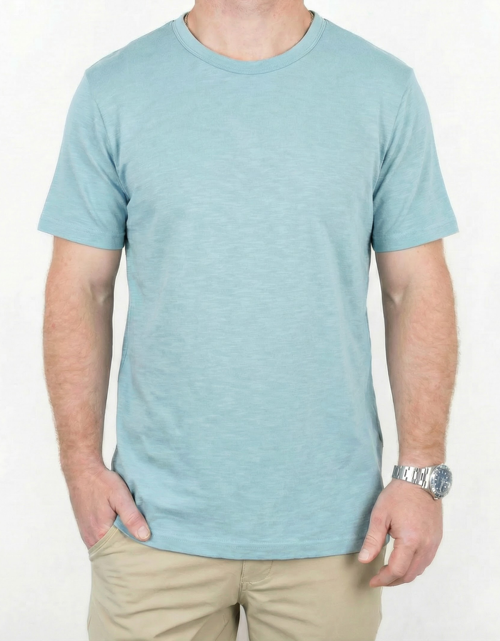 A close-up torso shot of a man wearing the light blue Baja Llama Solid Porcelain Blue Pima Slub Tee, featuring a distinct slub texture, styled with one hand in his khaki pants pocket and a silver watch