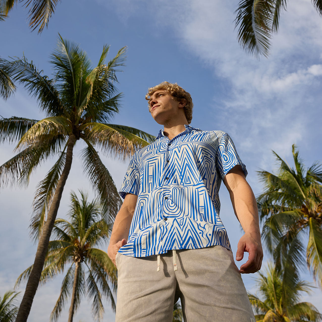 Man wearing Baja Llama Too Easys Acid Washed Gray Stretch Cotton Shorts and blue patterned button-up shirt under palm trees.