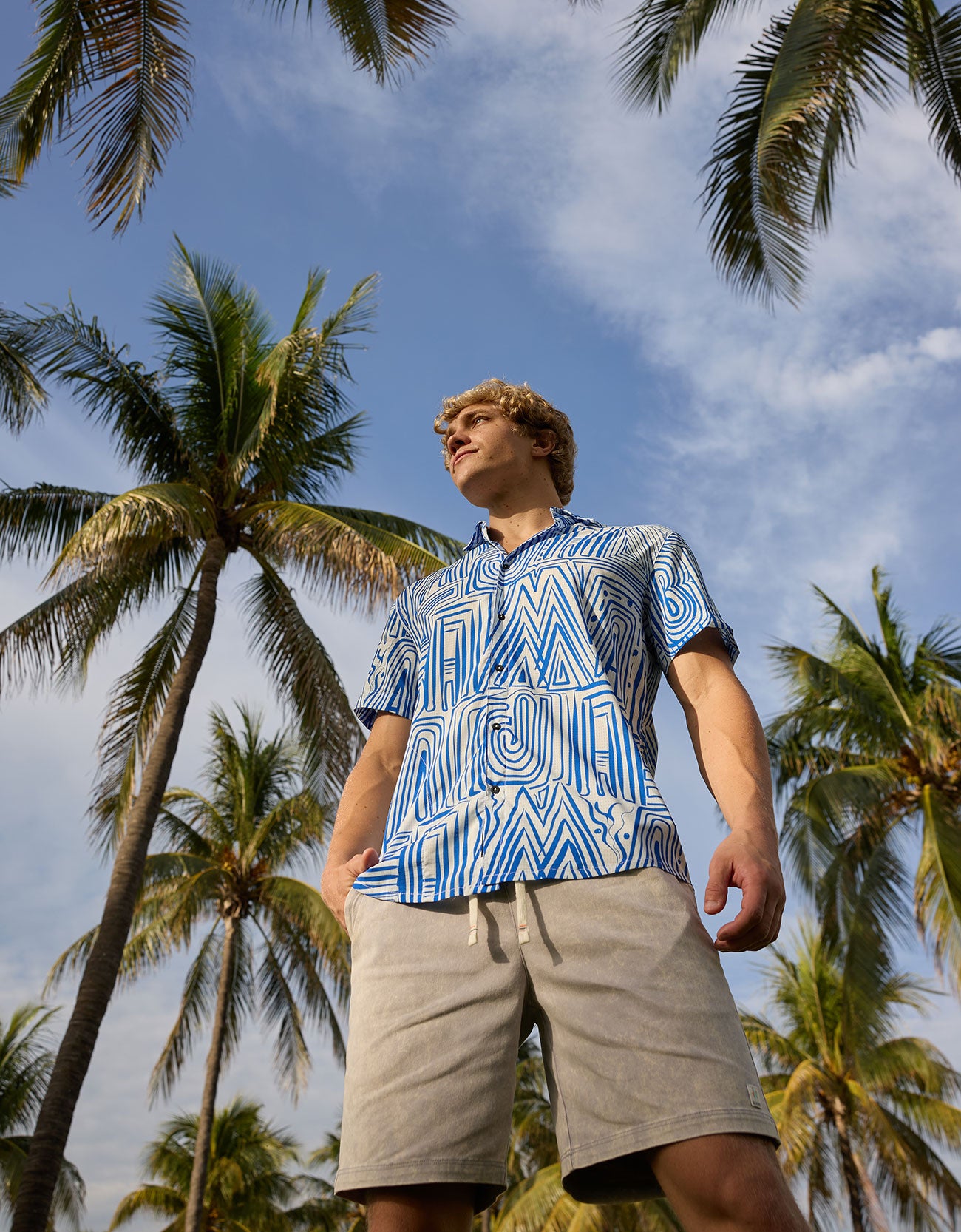 Man wearing Baja Llama Too Easys Acid Washed Gray Stretch Cotton Shorts and blue patterned button-up shirt under palm trees.