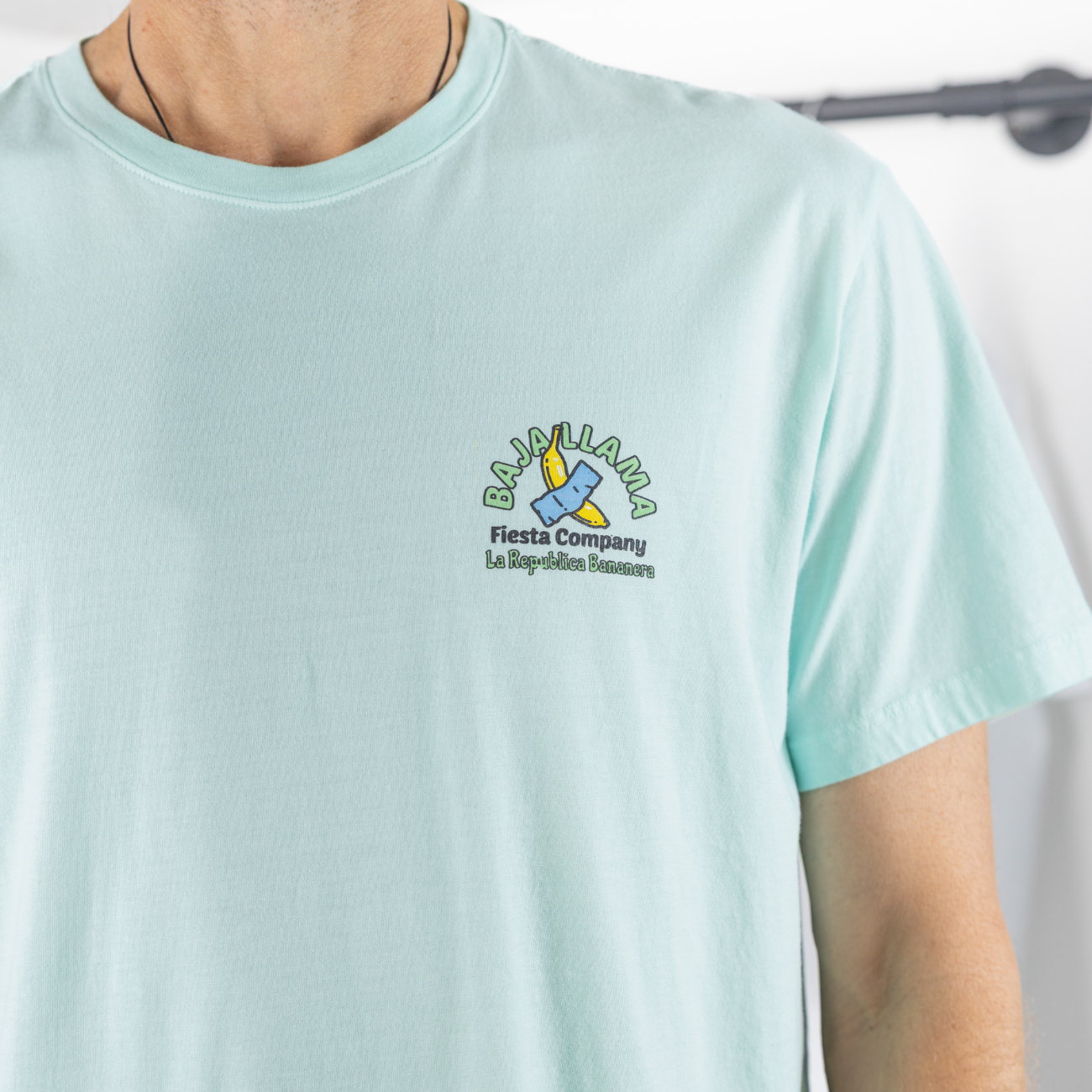 A close-up view of a model wearing the Baja Llama La Republica Bananera icy mint graphic tee, showcasing the small left-chest logo and crew neck design