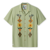 Front view of the Baja Llama 'Oasis Geometry' One-Off relaxed-fit short-sleeve button-up shirt in a soft sage green, featuring two vertical panels of symmetrical, colorful embroidery depicting palm trees, suns, and geometric diamond shapes.