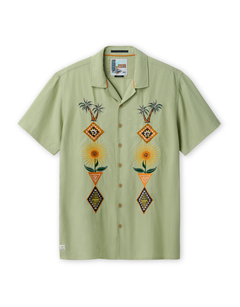 Front view of the Baja Llama 'Oasis Geometry' One-Off relaxed-fit short-sleeve button-up shirt in a soft sage green, featuring two vertical panels of symmetrical, colorful embroidery depicting palm trees, suns, and geometric diamond shapes.