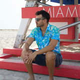 Model sitting near a Miami lifeguard stand wearing a Pastel Paradise reverse print blue and green perforated button-up shirt