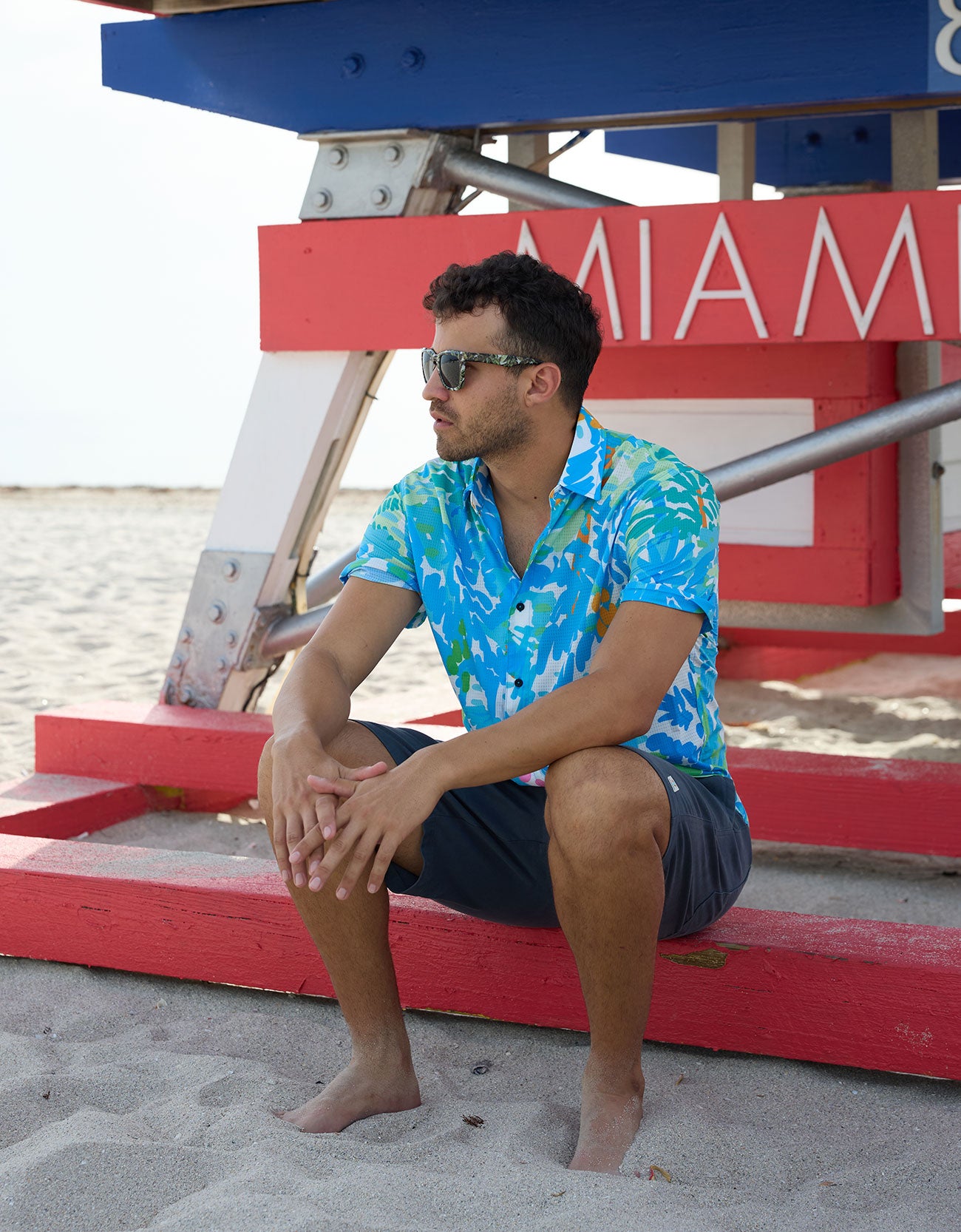 Model sitting near a Miami lifeguard stand wearing a Pastel Paradise reverse print blue and green perforated button-up shirt