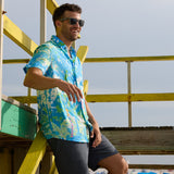 Model standing against a yellow beach structure wearing the Pastel Paradise reverse print blue and green perforated button-up shirt