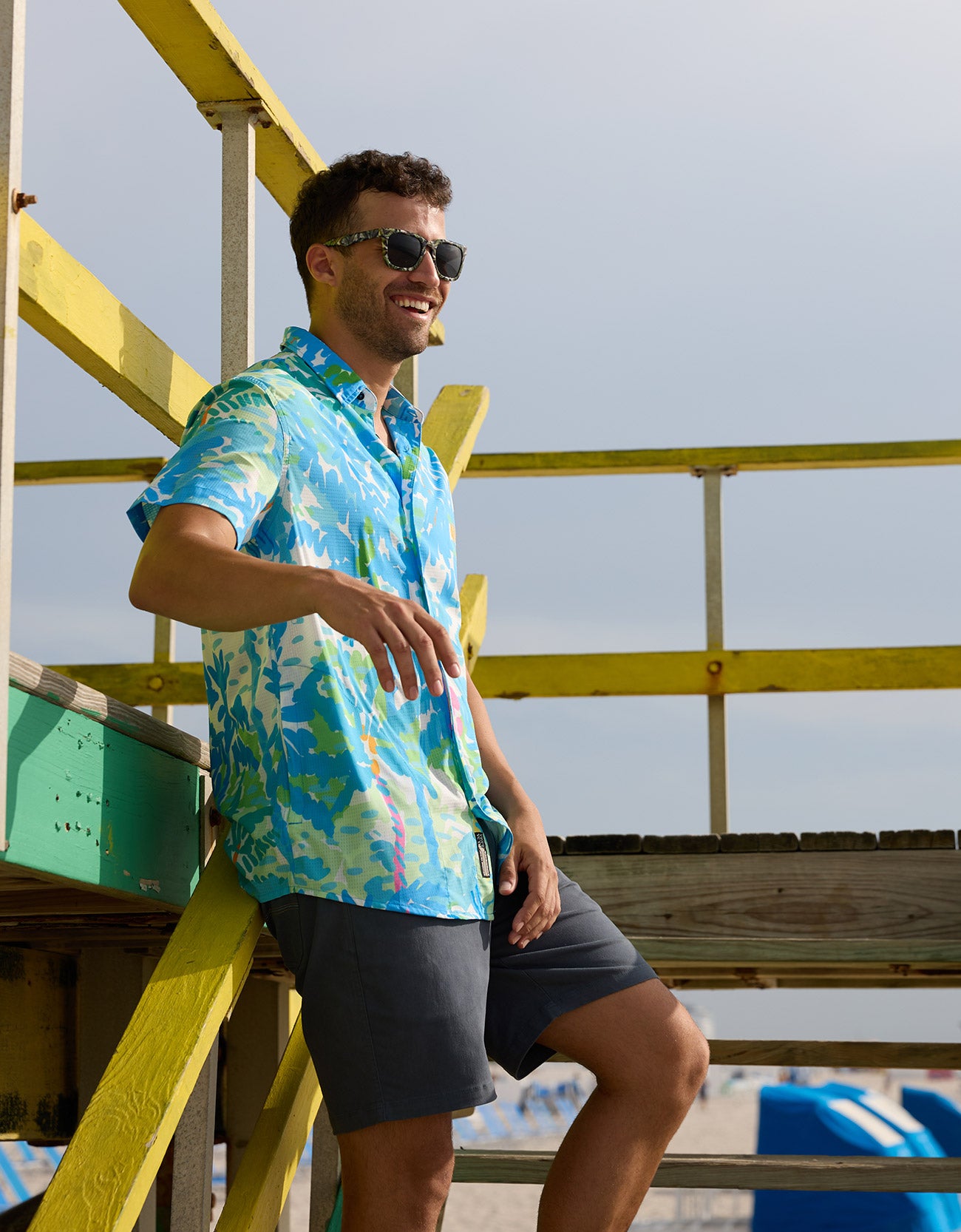 Model standing against a yellow beach structure wearing the Pastel Paradise reverse print blue and green perforated button-up shirt
