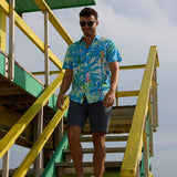 Model walking down green and yellow beach lifeguard tower stairs wearing Pastel Paradise reverse print blue perforated short-sleeve shirt