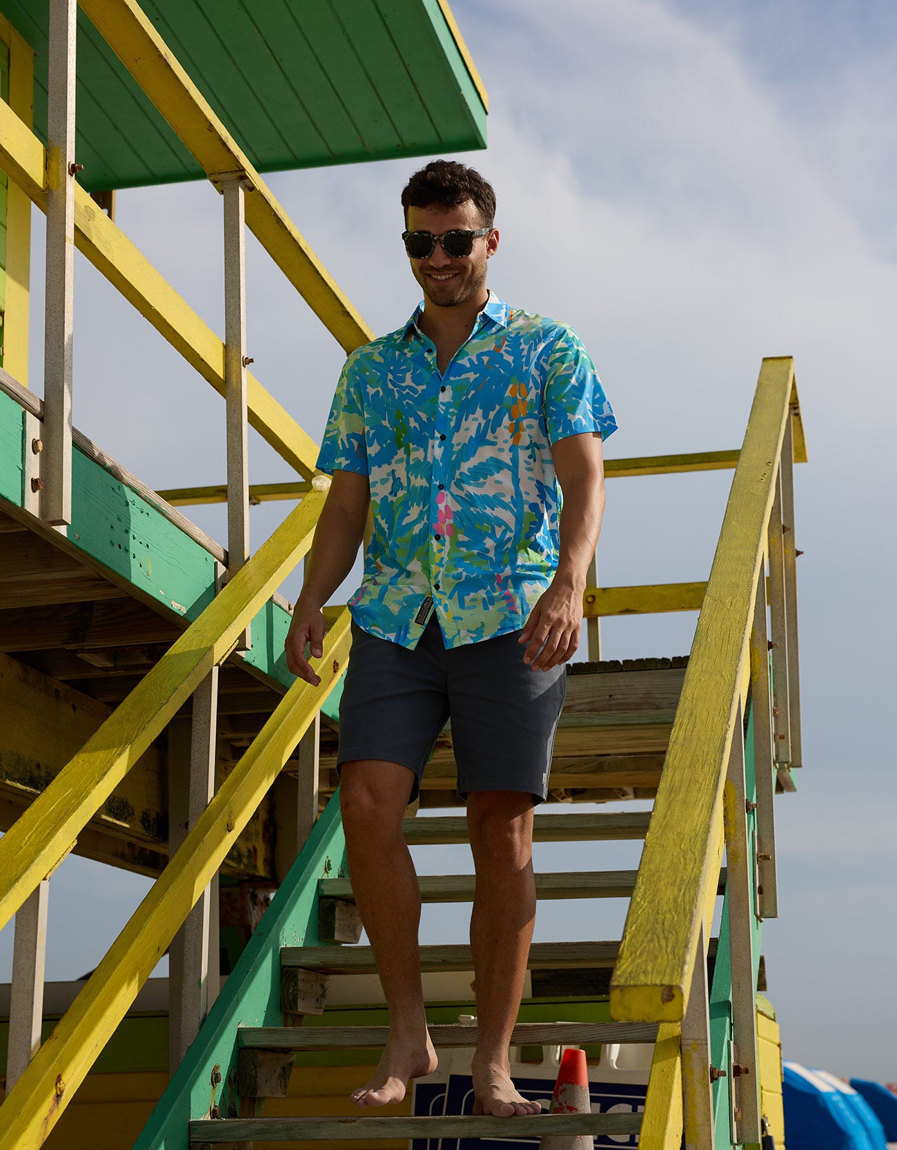 Model walking down green and yellow beach lifeguard tower stairs wearing Pastel Paradise reverse print blue perforated short-sleeve shirt