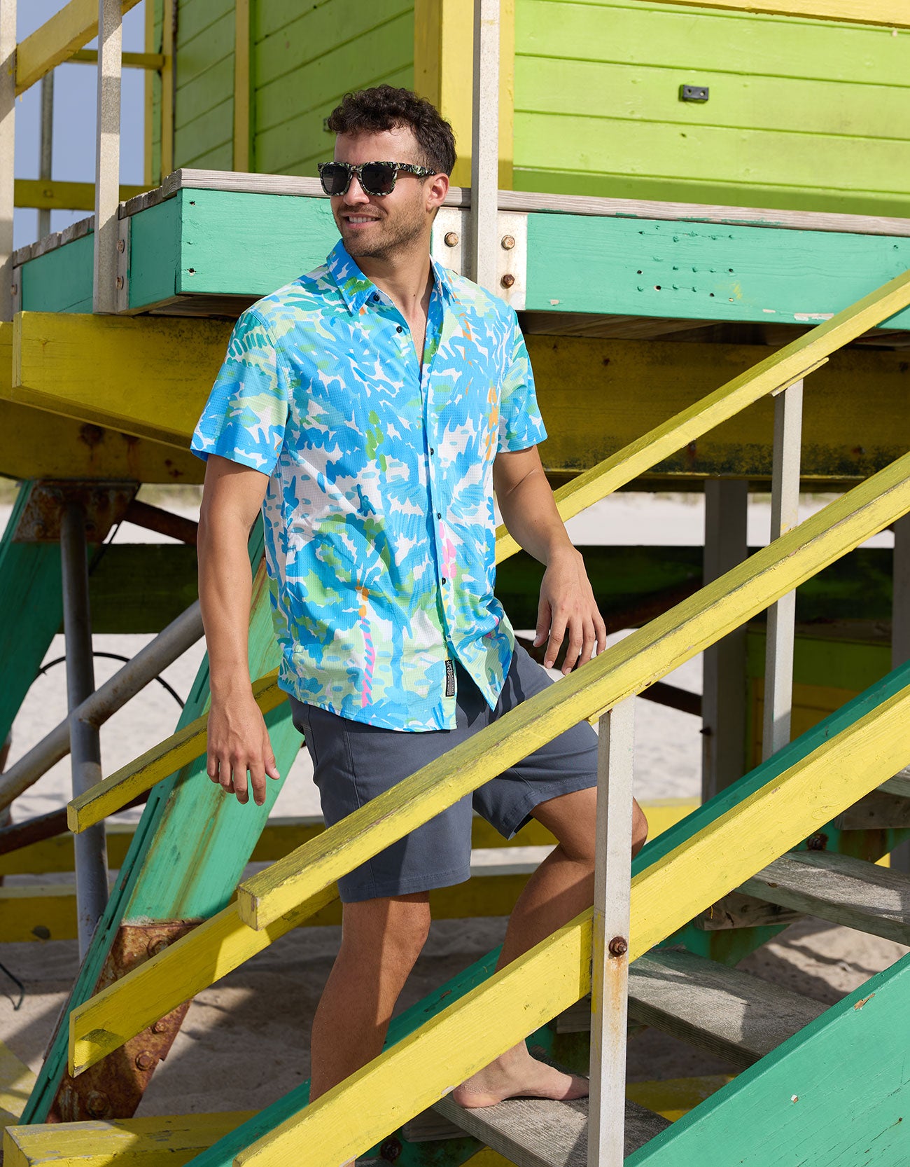 Model standing on green and yellow beach lifeguard tower stairs wearing Pastel Paradise reverse print blue and green perforated button-up shirt