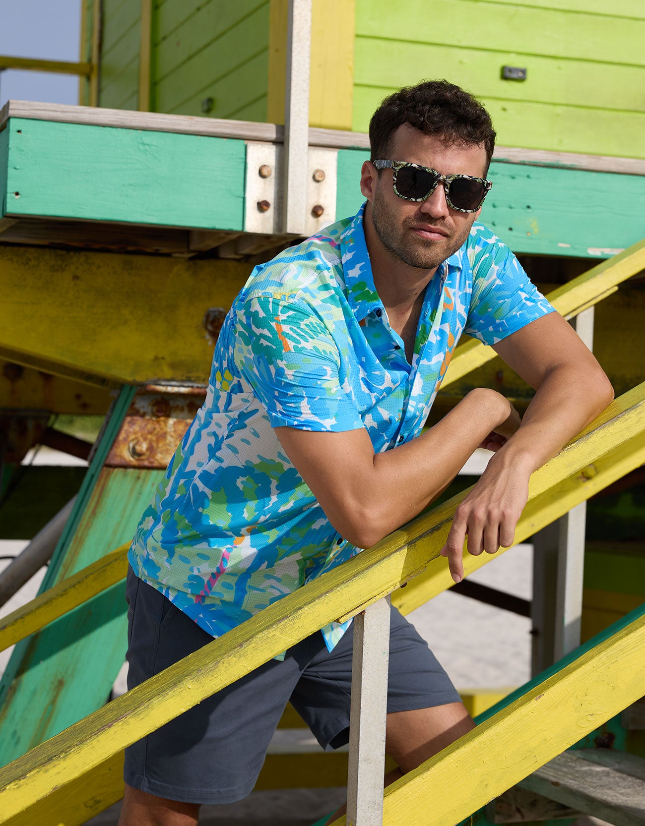 Model leaning on yellow lifeguard stairs wearing Pastel Paradise reverse print blue and green perforated button-up shirt and sunglasses