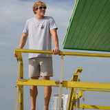 Model standing on green and yellow lifeguard tower wearing the Radical Misadventurist purple acid wash graphic tee and sunglasses