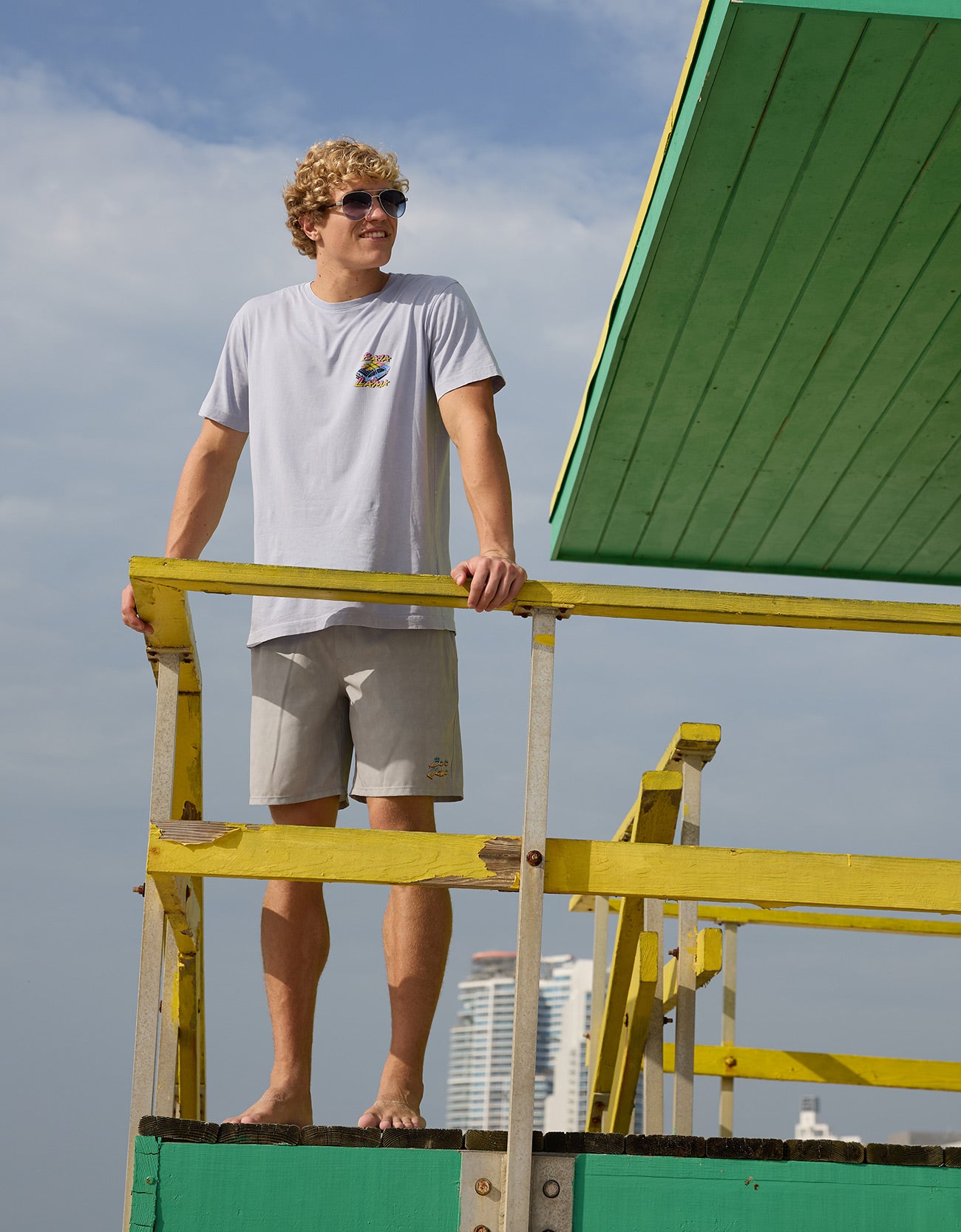 Model standing on green and yellow lifeguard tower wearing the Radical Misadventurist purple acid wash graphic tee and sunglasses