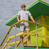 Model standing on green and yellow lifeguard tower stairs wearing the Radical Misadventurist purple acid wash graphic tee and sunglasses