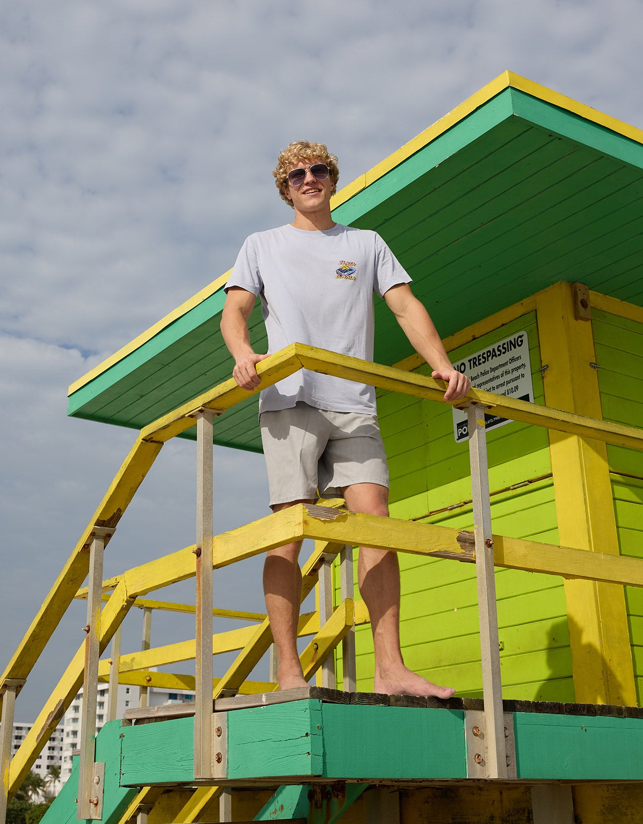 Model standing on green and yellow lifeguard tower stairs wearing the Radical Misadventurist purple acid wash graphic tee and sunglasses
