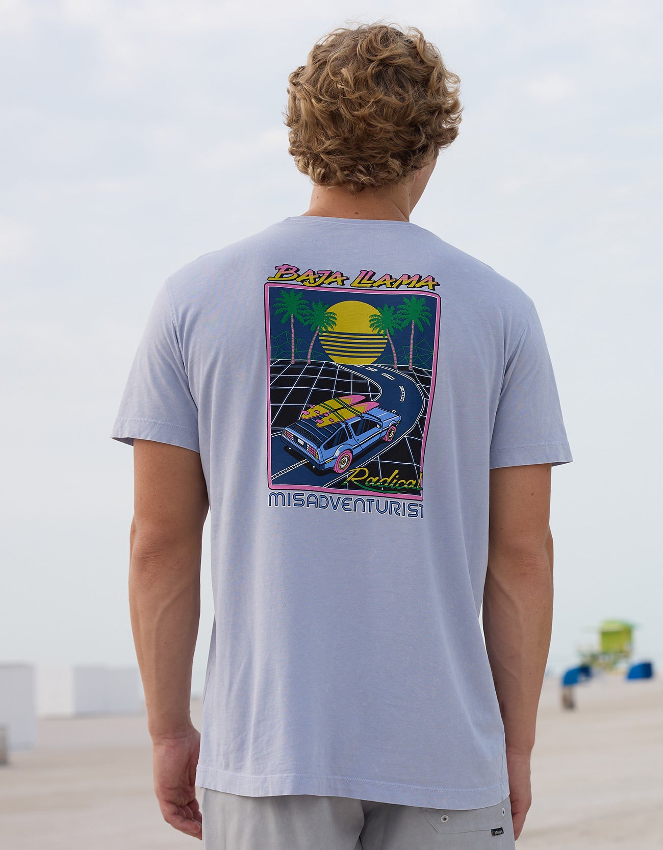 Back view of a model wearing the Radical Misadventurist purple acid wash graphic tee featuring a retro car and palm tree back print