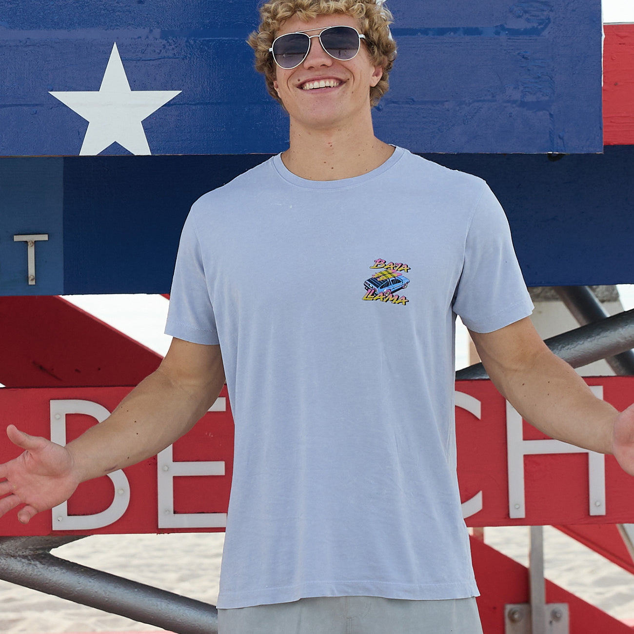 Model wearing Baja Llama Radical Misadventurist purple acid wash graphic tee with sunglasses in front of a red, white, and blue lifeguard tower