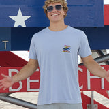 Model wearing Baja Llama Radical Misadventurist purple acid wash graphic tee with sunglasses in front of a red, white, and blue lifeguard tower