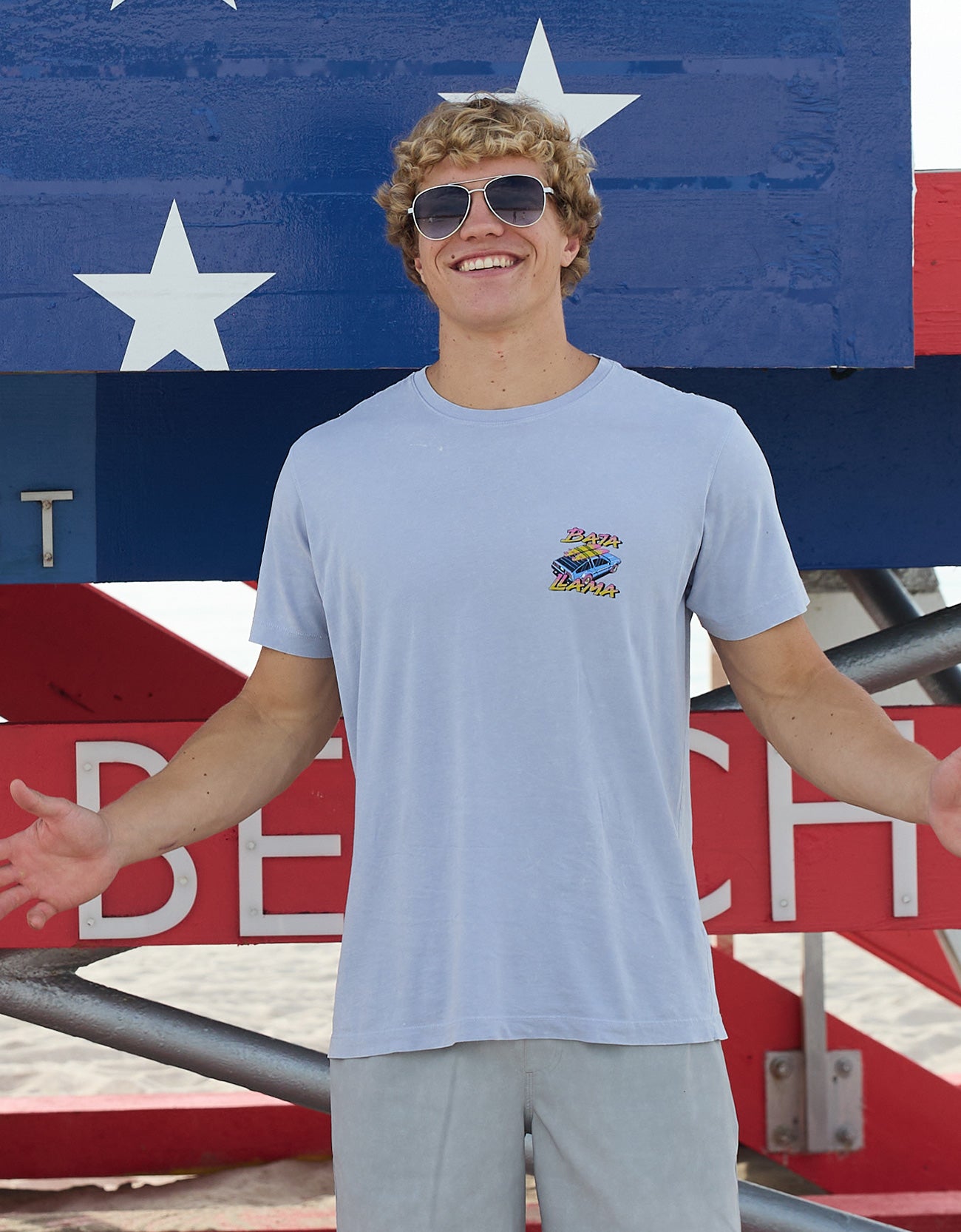 Model wearing Baja Llama Radical Misadventurist purple acid wash graphic tee with sunglasses in front of a red, white, and blue lifeguard tower