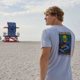 Model looking toward an American flag lifeguard tower on the beach wearing the Radical Misadventurist purple acid wash graphic tee showing back print