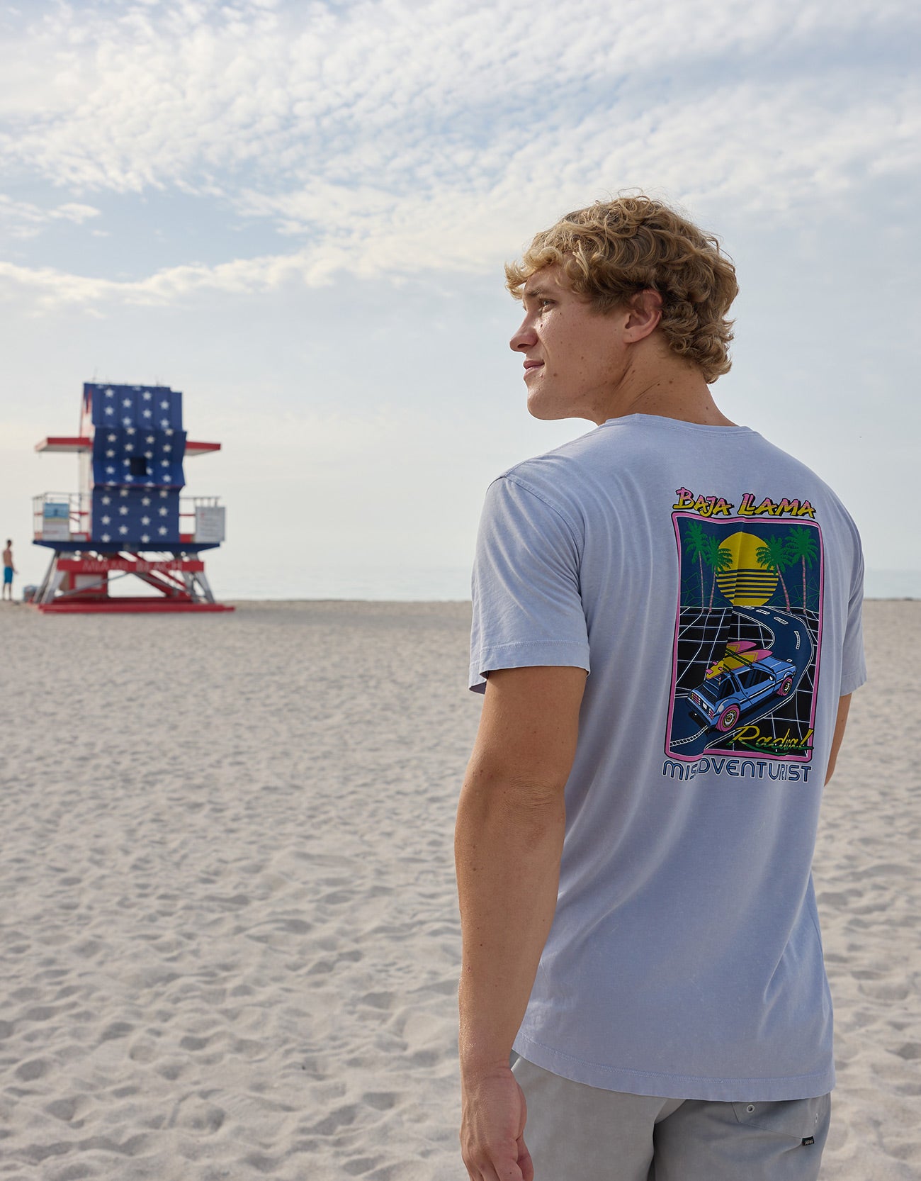 Model looking toward an American flag lifeguard tower on the beach wearing the Radical Misadventurist purple acid wash graphic tee showing back print